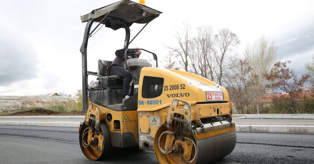 A road roller compacting fresh asphalt on a new roadway with a construction worker operating the vehicle.
