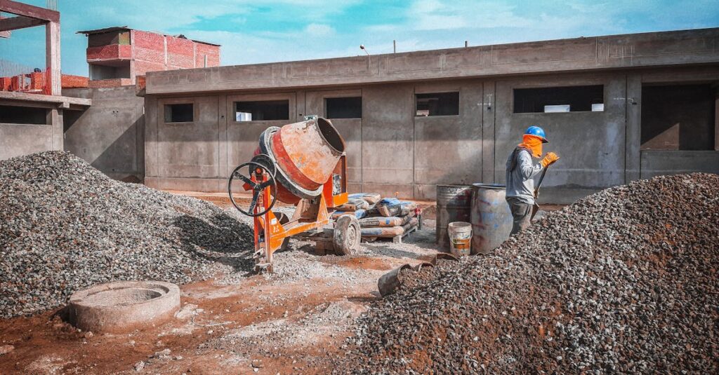 A construction site with a worker wearing safety gear next to a cement mixer and gravel piles.